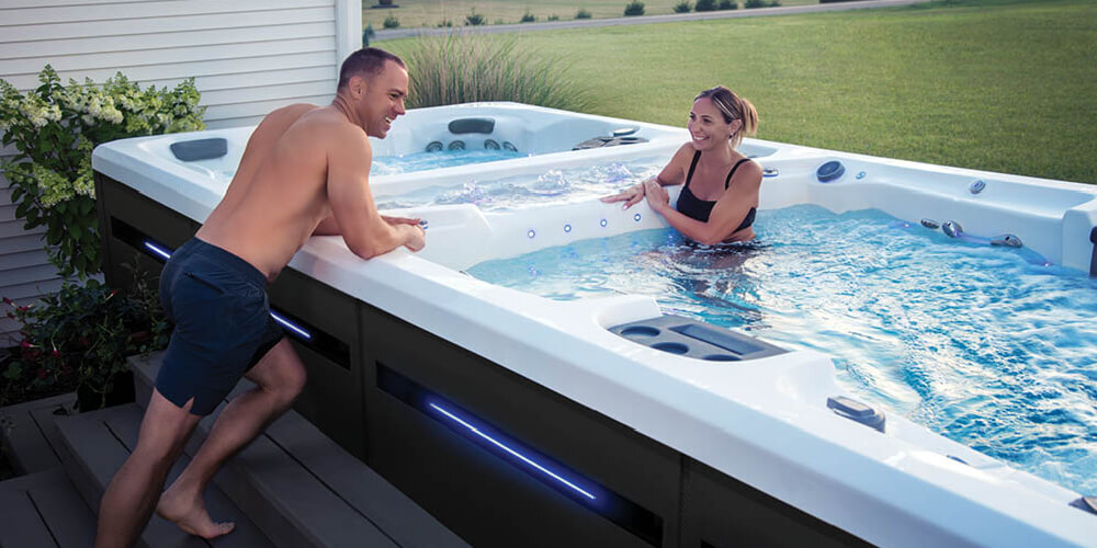 Un homme appuyé contre le bord d'un spa de nage, en train de parler à une femme qui se trouve dans l'eau.